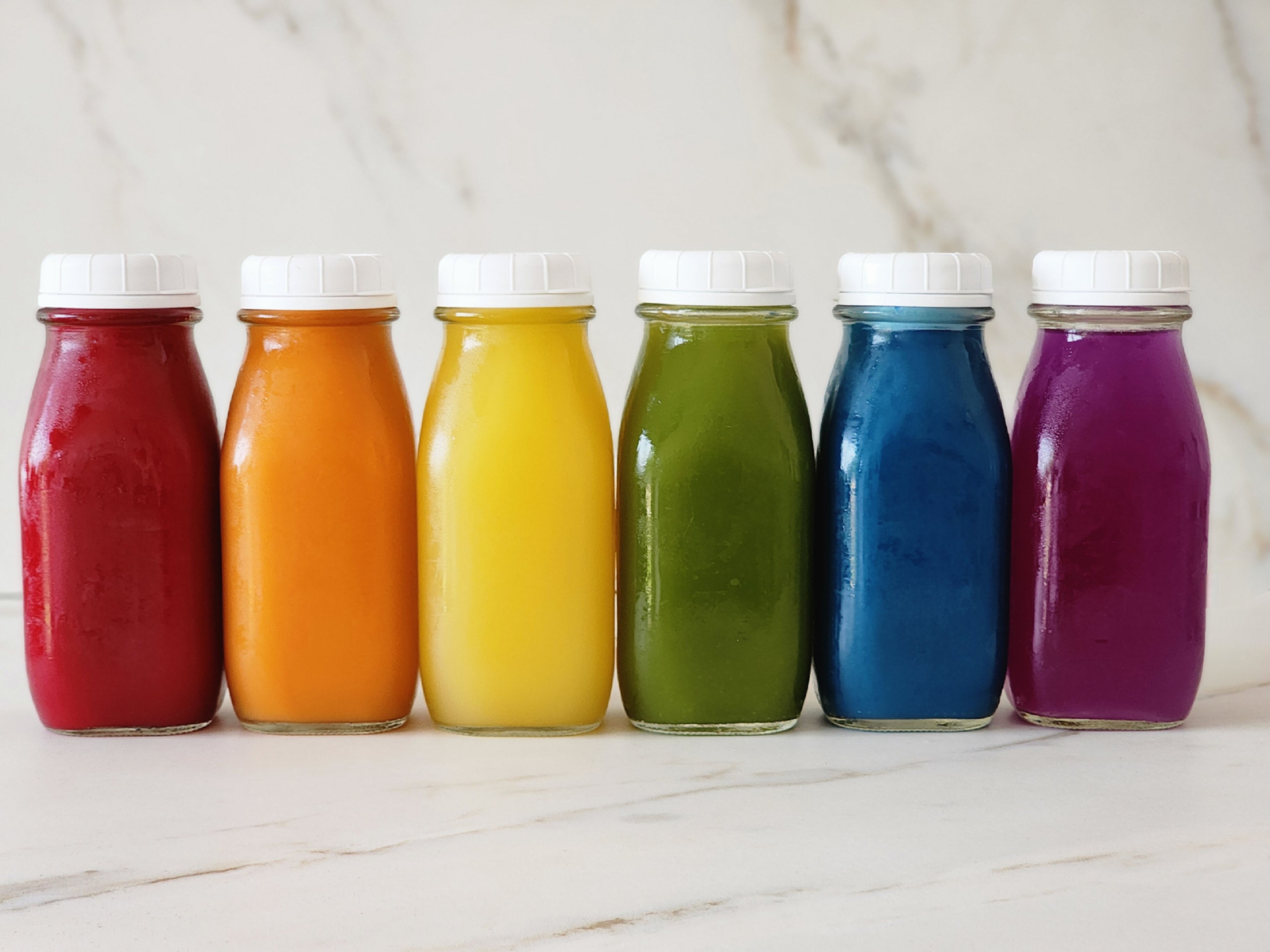 Photo of cold-press juices in glass bottles with white bottle caps in a row. Colorful juices red, orange, yellow, green, blue and purple.